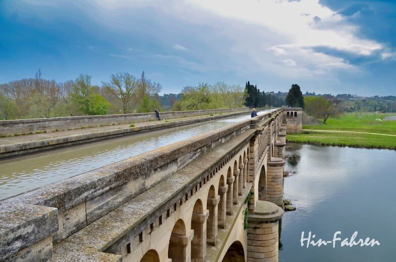 Eine lange Bogenbrücke aus Stein führt über einen Fluss, der sowohl einen Kanal als auch einen Weg führt. Bäume und grasbewachsene Felder säumen die Ufer unter einem bewölkten blauen Himmel. In der unteren rechten Ecke stehen die Worte Hin-Fahren.