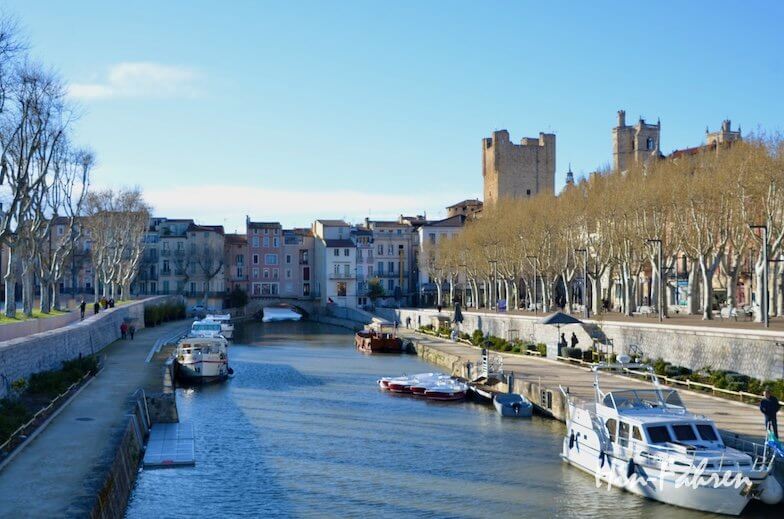 Ein mit Booten gesäumter Kanal führt durch eine Stadt mit pastellfarbenen Gebäuden und blattlosen Bäumen. Auf beiden Seiten des Kanals gehen Menschen spazieren, und im Hintergrund erhebt sich ein historischer Steinturm unter einem klaren blauen Himmel.