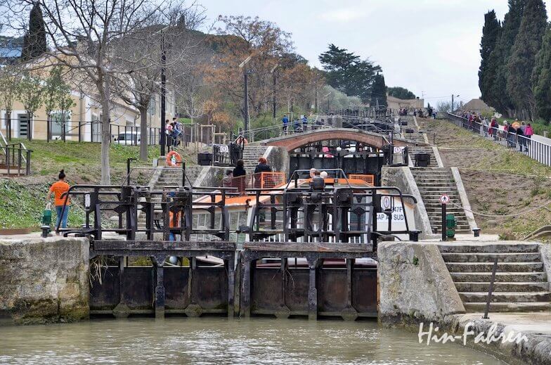 Eine Kanalschleusenanlage mit mehreren Toren und Booten, umgeben von Spaziergängern und Beobachtern. Bäume und Gebäude säumen die Seiten, und die Atmosphäre ist lebhaft und geschäftig.