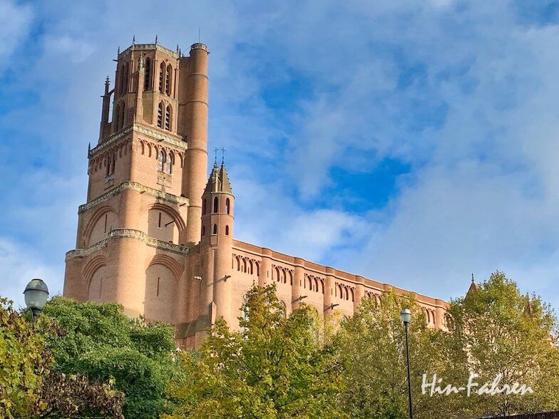 Eine große Backsteinkathedrale mit hohen Türmen steht vor einem blauen Himmel mit vereinzelten Wolken, umgeben von grünen Bäumen.