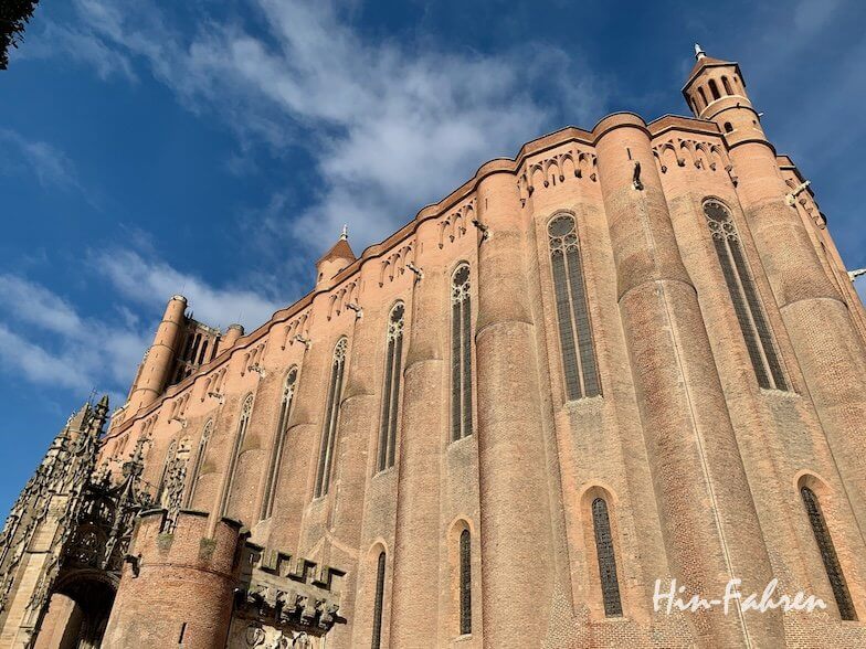 Blick aus geringer Höhe auf eine große gotische Kathedrale aus rotem Backstein mit hohen vertikalen Fenstern und komplizierten Details vor einem blauen Himmel mit vereinzelten Wolken. Die Worte Hin-Fahren erscheinen in der unteren rechten Ecke.
