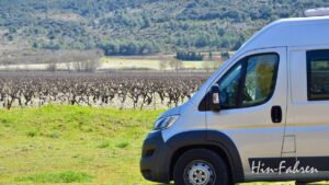 Ein silbernes Wohnmobil parkt auf einer Wiese in der Nähe eines Weinbergs mit kahlen Rebstöcken; im Hintergrund sind grüne Hügel und Bäume unter einem klaren Himmel zu sehen.