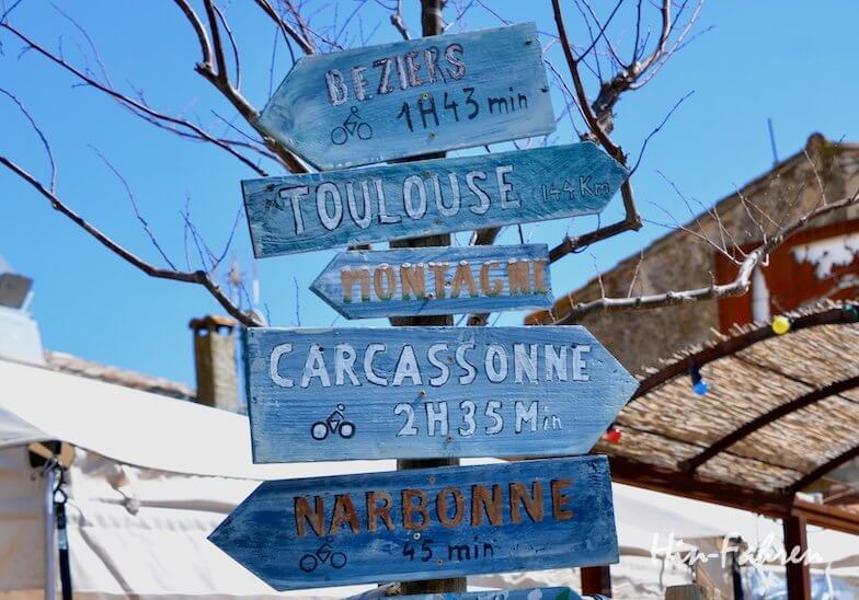 Vier blaue Holzschilder mit handgeschriebenen Entfernungsangaben weisen nach Béziers, Toulouse, Carcassonne und Narbonne, mit kleinen Fahrradsymbolen, vor einem Hintergrund aus kahlen Ästen und einem rustikalen Gebäude.