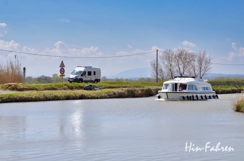 Ein weißes Wohnmobil steht auf einer grasbewachsenen Straße an einem Kanal, während ein weißes Kanalboot unter einem klaren blauen Himmel mit vereinzelten Wolken auf dem Wasser fährt.
