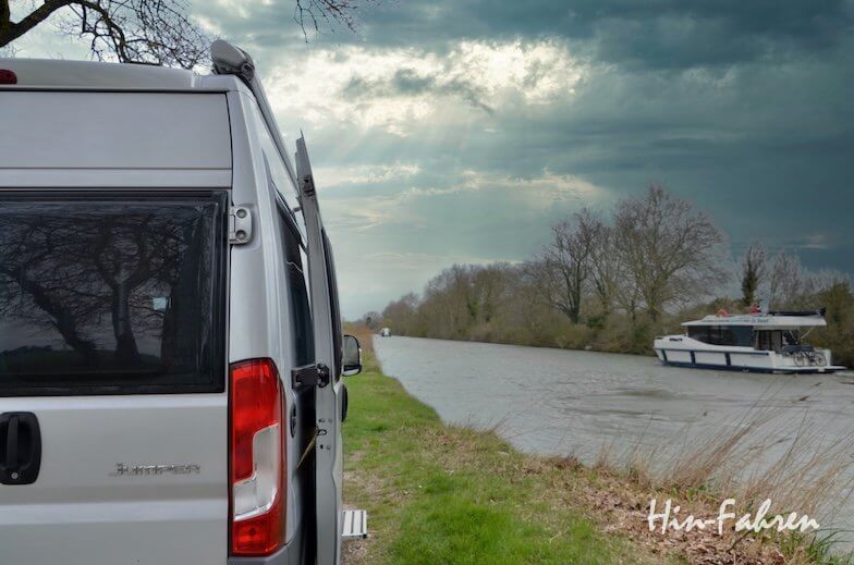 Ein Wohnmobil parkt an einem ruhigen Kanal mit einem Boot auf dem Wasser. Bäume säumen beide Seiten des Kanals, und der Himmel ist bewölkt mit durchbrechenden Sonnenstrahlen. In der unteren rechten Ecke erscheint der Text "HinFahren".