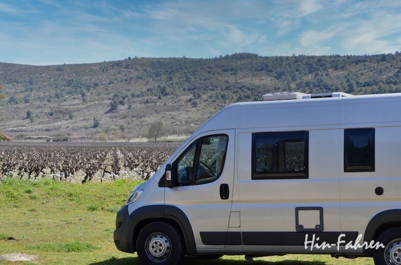 Ein silbernes Wohnmobil ist auf einer Wiese neben einem Weinberg mit Weinreben geparkt, vor dem Hintergrund grüner Hügel unter blauem Himmel. In der rechten unteren Ecke ist das Wort Hin-Fahren zu lesen.