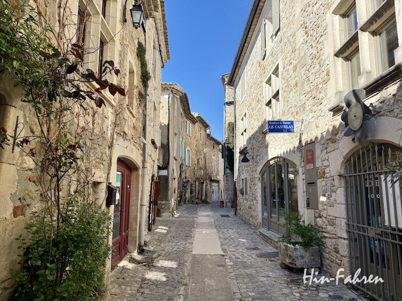 Eine schmale Straße mit Kopfsteinpflaster und alten Steinhäusern schlängelt sich durch ein historisches Ardeche-Dorf, das unter einem strahlend blauen Himmel im Sonnenlicht liegt.