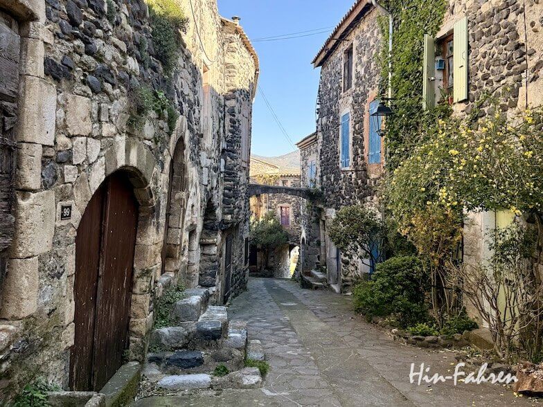 Eine schmale Steinstraße schlängelt sich zwischen alten Steinhäusern mit Holztüren und grün beschlagenen Fenstern, die von Pflanzen gesäumt und von der Sonne beschienen werden, durch ein ruhiges, historisches Ardeche-Dorf.