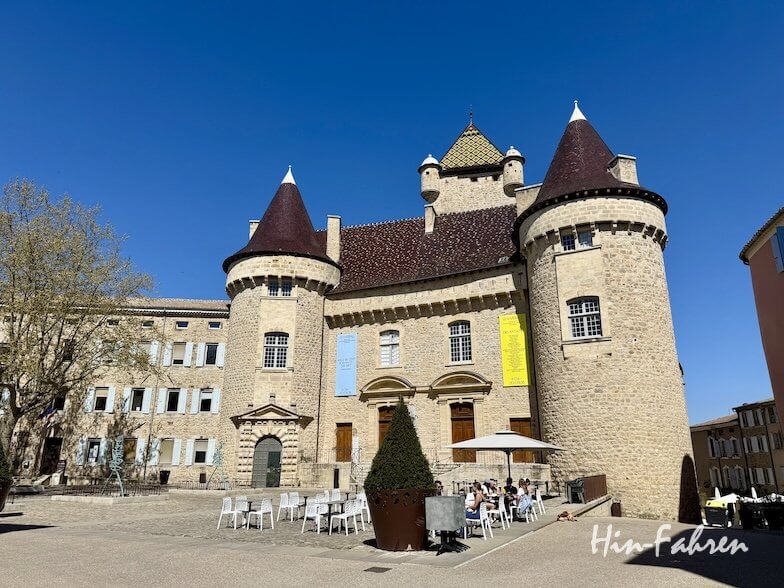 Ein großes Steinschloss mit runden Türmen und kegelförmigen Dächern steht auf einem sonnigen Platz an der Ardeche. Davor sitzen Menschen an Tischen im Freien, und in der Nähe des Eingangs hängen Transparente. Der Himmel ist klar und blau.