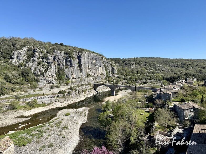 Eine Steinbrücke überspannt einen flachen Fluss mit felsigen Ufern in der Ardeche, umgeben von Felsen und grünen Hügeln. Traditionelle Steinhäuser stehen in der Nähe des Wassers unter einem klaren blauen Himmel.