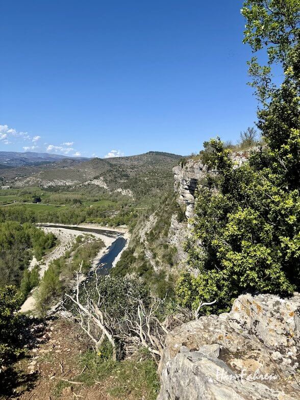 Ein malerischer Blick auf den gewundenen Fluss Ardeche, der sich durch ein grünes Tal schlängelt, mit felsigen Klippen und Hügeln im Hintergrund unter einem klaren blauen Himmel. Im Vordergrund sind Bäume und Sträucher zu sehen.