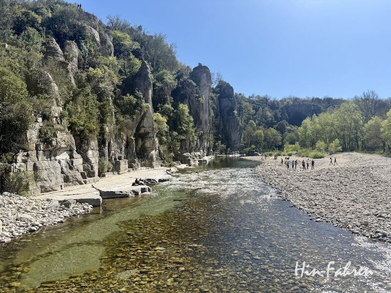 Ein klarer, flacher Fluss fließt neben felsigen Klippen und einem Kieselufer in der Ardèche, umgeben von grünen Bäumen unter einem blauen Himmel. Kleine Gruppen von Menschen spazieren am Flussufer entlang.