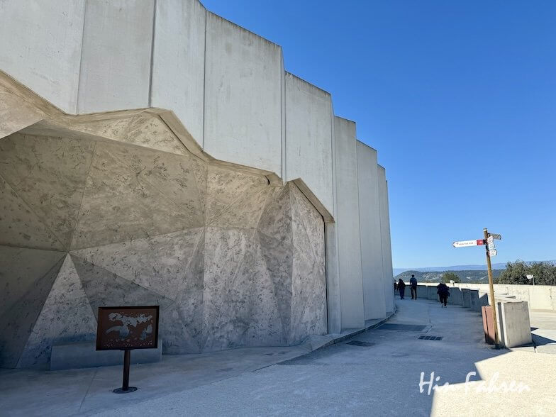 Eine moderne, kantige Betonstruktur mit geometrischen Mustern steht neben einem Weg in der Ardeche. Ein Wegweiser und eine Informationstafel stehen in der Nähe, während mehrere Personen bei strahlend blauem Himmel den Weg entlanggehen.