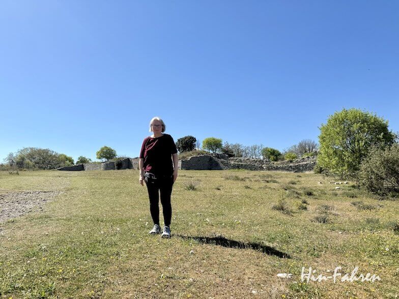 Eine Person mit kurzen, hellen Haaren steht auf einer grasbewachsenen Wiese unter einem klaren, blauen Himmel in der Ardeche, mit verstreuten Bäumen und Steinruinen im Hintergrund.