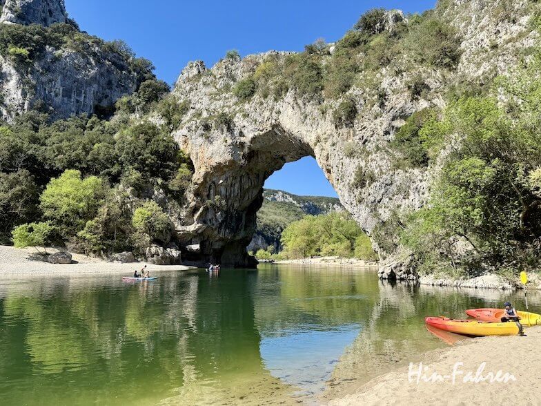 Ein ruhiger Fluss fließt unter einem großen natürlichen Steinbogen in der Ardeche, umgeben von felsigen Klippen und grünen Bäumen. Die Menschen fahren Kajak und entspannen sich an einem sonnigen Tag am Wasser. Hin-Fahren steht in weißer Schrift unten rechts.