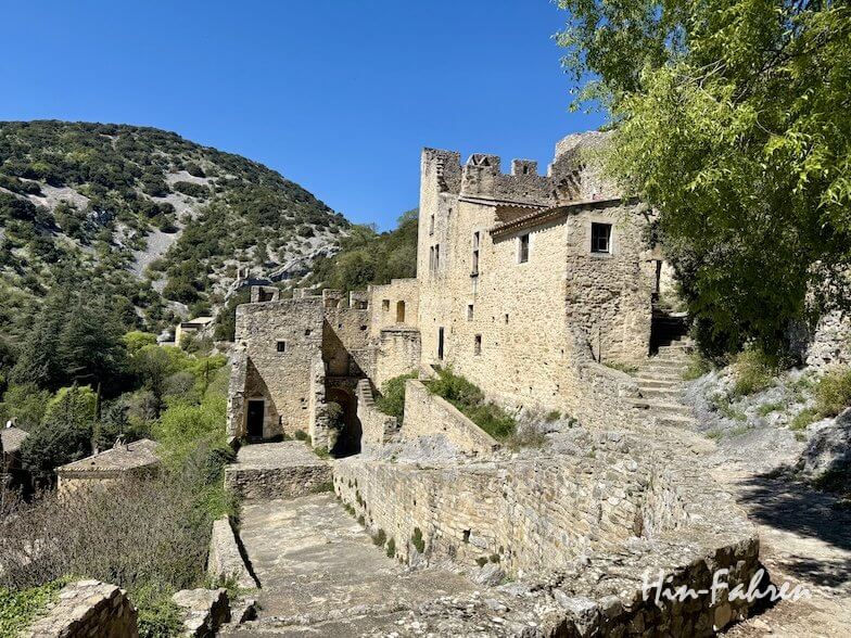 Eine alte steinerne Festung mit gewölbten Türen und kleinen Fenstern liegt auf einem Hügel in der Ardeche, umgeben von grünen Bäumen und Sträuchern, mit einer Berglandschaft im Hintergrund unter einem klaren blauen Himmel.