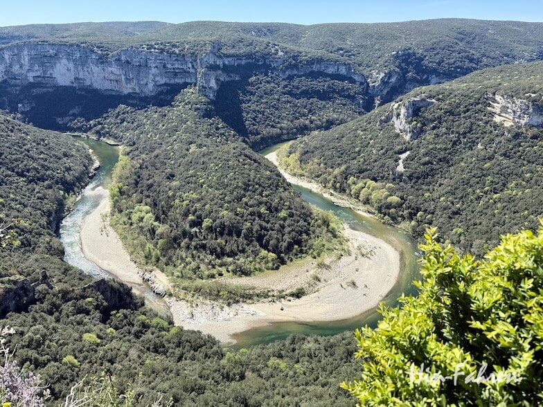 Ein sich schlängelnder Fluss schlängelt sich durch ein üppiges, grünes Ardeche-Tal, das von steilen, felsigen Hügeln und Klippen umgeben ist, unter einem klaren blauen Himmel. Eine dichte Vegetation bedeckt die Landschaft und hebt die dramatische Flussbiegung hervor.