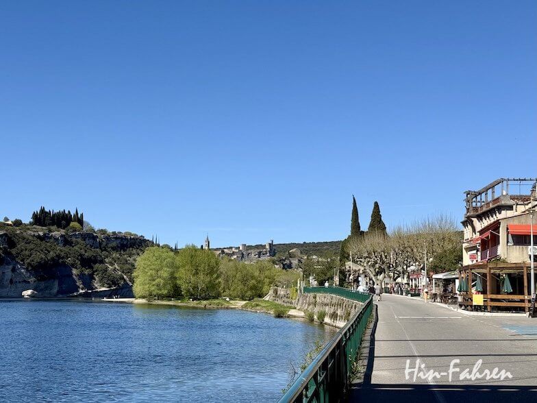 Eine malerische Uferstraße in der Ardeche mit grünen Bäumen, einem ruhigen blauen Fluss und farbenfrohen Gebäuden unter einem klaren blauen Himmel; in der Ferne sind Hügel und ein Dorf zu sehen.