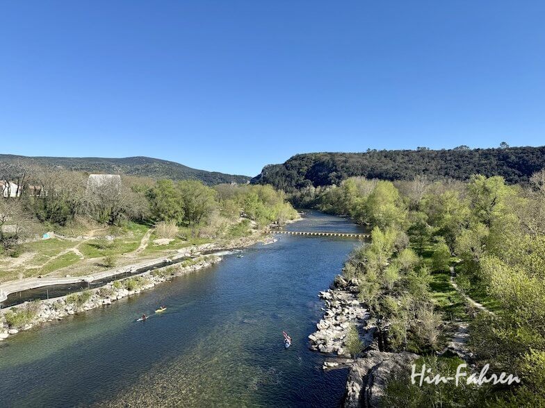 Ein klarer Fluss schlängelt sich unter strahlend blauem Himmel durch die üppige, von Bäumen gesäumte Landschaft der Ardeche, mit Hügeln in der Ferne und einigen Kajak- oder Kanufahrern. Der Text Hin-Fahren erscheint in der unteren rechten Ecke.