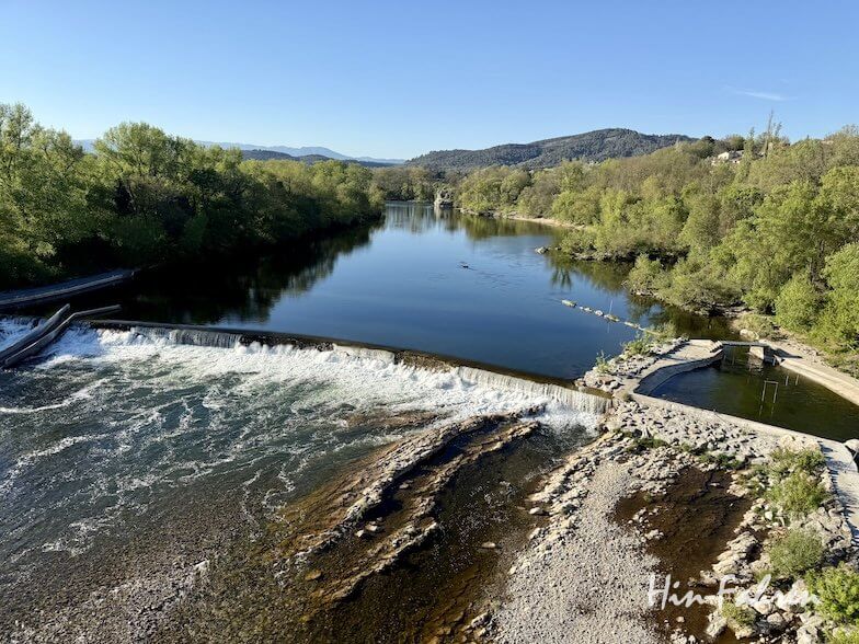 Ein ruhiger Fluss fließt über ein niedriges Betonwehr in der Ardeche, umgeben von üppigen grünen Bäumen, mit felsigen Ufern und fernen Hügeln unter einem klaren blauen Himmel.