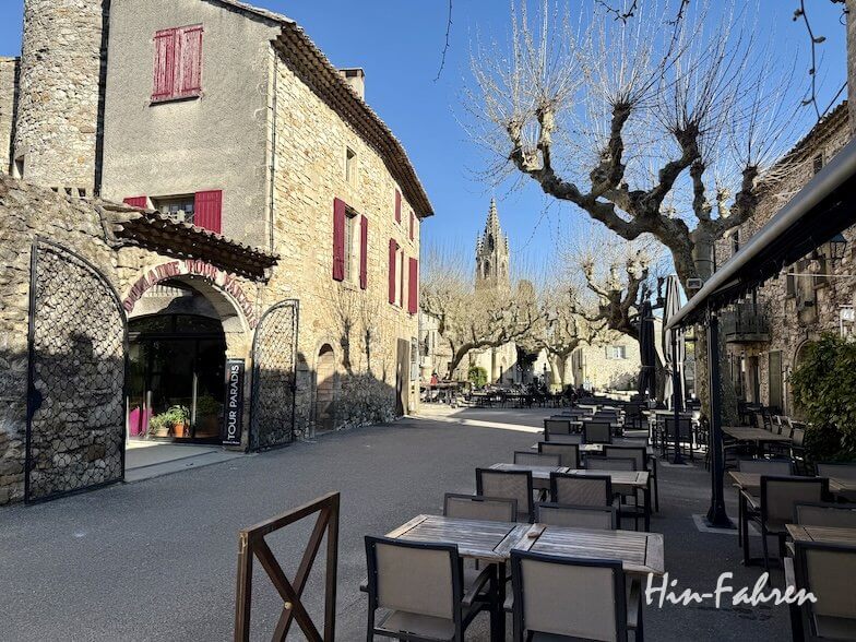 Eine ruhige Kopfsteinpflasterstraße in einem Dorf an der Ardeche, gesäumt von Tischen im Freien, Steinhäusern mit roten Fensterläden, blattlosen Bäumen und einem Kirchturm, der sich unter einem klaren blauen Himmel erhebt.