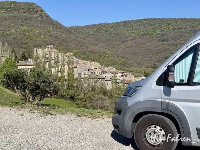 Ein silbernes Wohnmobil, geparkt auf einem Schotterplatz, überblickt ein Ardeche-Dorf mit Steinhäusern, umgeben von grünen Bäumen und bewaldeten Bergen unter einem klaren blauen Himmel.