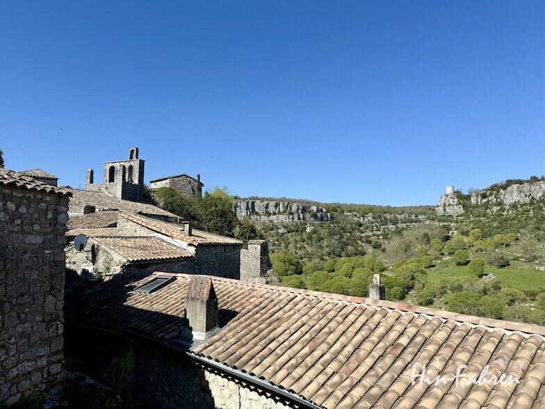Steinhäuser mit Terrakotta-Dächern stehen an einem Hang unter blauem Himmel, umgeben von grünen Bäumen und Felsen im Hintergrund, die den Charme eines historischen Ardeche-Dorfes in einer ländlichen, bergigen Gegend ausstrahlen.