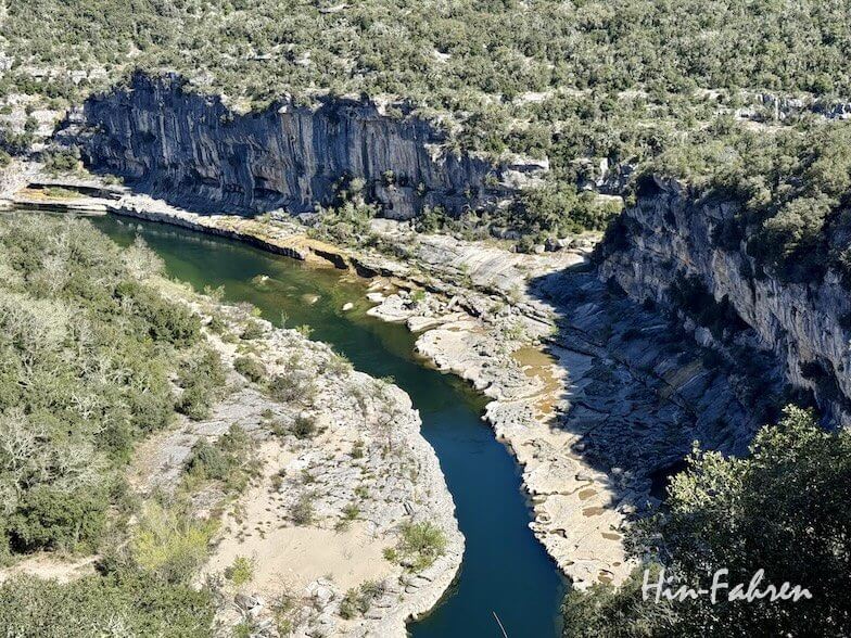 Ein gewundener Fluss fließt zwischen felsigen Klippen und dichten Ardeche-Wäldern im hellen Sonnenlicht, wobei die Schatten die zerklüftete Landschaft betonen. In der rechten unteren Ecke steht der Text "Hun-Fahren".