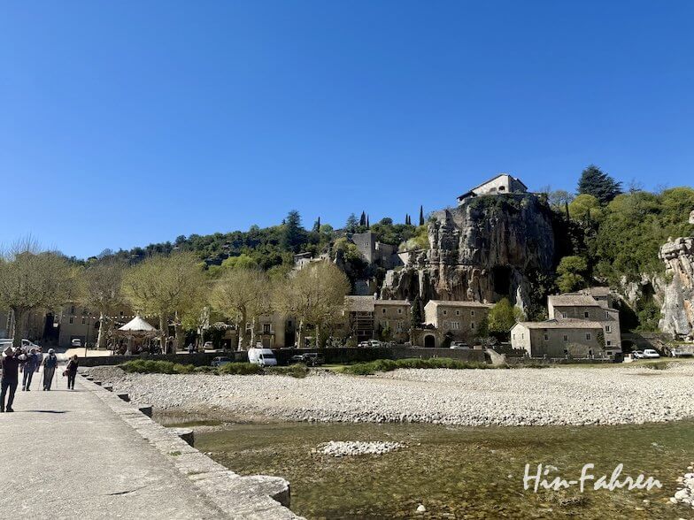 Ein Dorf am Flussufer der Ardeche mit steinernen Gebäuden, die sich an eine felsige Klippe schmiegen, umgeben von grünen Bäumen und Hügeln unter einem klaren blauen Himmel. Menschen spazieren entlang des Flusses und ein großes Haus thront auf dem Felsen.