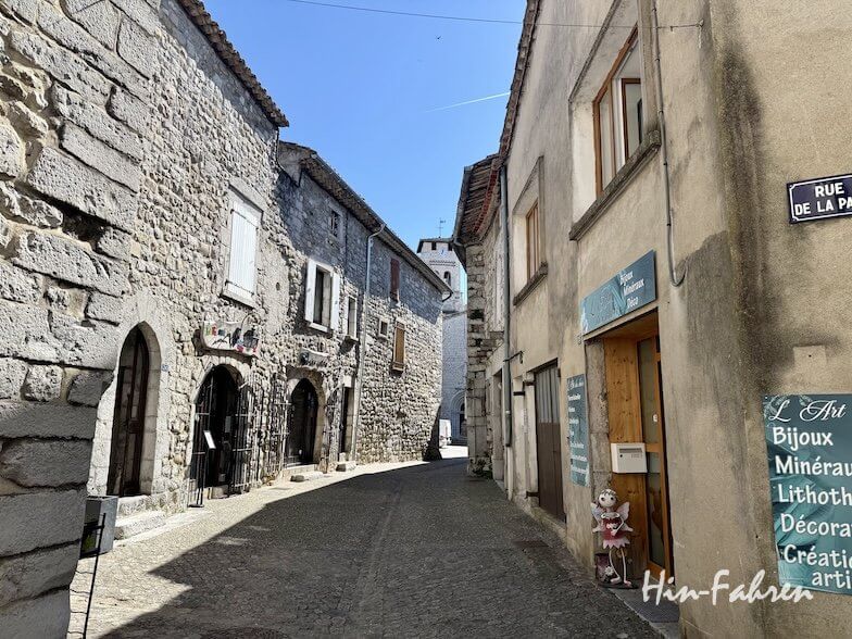 Schmale Kopfsteinpflasterstraße in einem historischen Dorf der Ardeche, gesäumt von Steinhäusern und Geschäften. Auf der rechten Seite steht ein blaues Straßenschild, und das Sonnenlicht wirft Schatten auf die Mauern und den Boden.