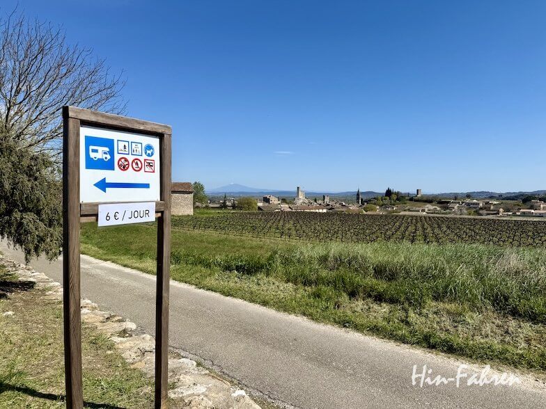 Ein Schild an einer Landstraße der Ardeche weist links auf einen Wohnmobilstellplatz für 6 Euro pro Tag hin, mit Symbolen für Parken, Wasser und Abfallentsorgung. Unter dem strahlend blauen Himmel sind Weinberge und ein Dorf mit einem Turm zu sehen.