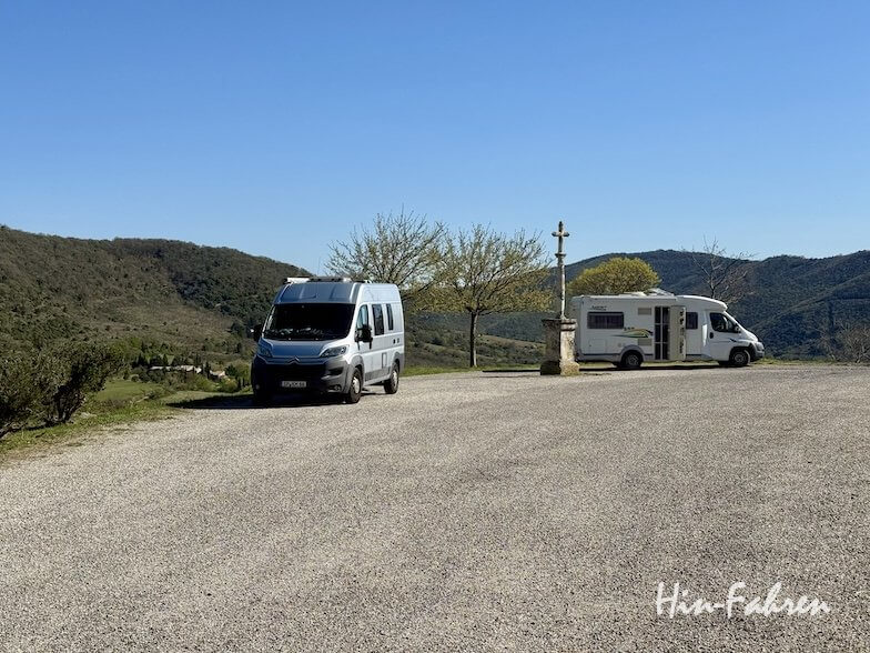 Zwei Wohnmobile stehen auf einem Schotterplatz in der Ardèche. Im Hintergrund sind Hügel, Bäume und ein strahlend blauer Himmel zu sehen. Zwischen den Fahrzeugen steht ein Steinmonument. Die Szene ist friedlich und sonnig.