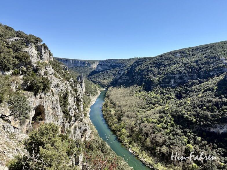 Ein sich schlängelnder Fluss fließt zwischen steilen, felsigen Klippen, die mit grüner Vegetation bedeckt sind, unter einem klaren blauen Himmel und bietet einen malerischen Blick auf das Ardeche-Tal.