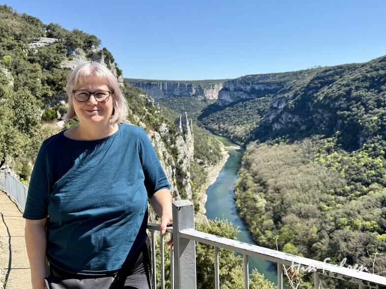 Eine Frau mit grauem Haar und Brille steht an einem Geländer und blickt auf die tiefe Schlucht des Flusses Ardeche, umrahmt von grünen Hügeln und einem klaren blauen Himmel im Hintergrund.
