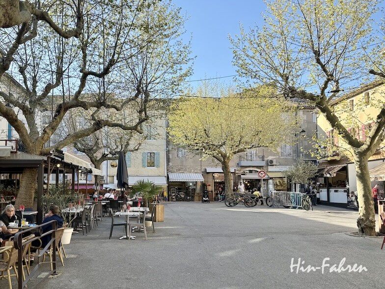 Ein sonniger Platz in der Ardeche mit blühenden Bäumen, Menschen, die sich an Cafétischen ausruhen, Fahrrädern, die in der Nähe geparkt sind, und mit Geschäften gesäumten Gebäuden im Hintergrund unter einem klaren blauen Himmel.