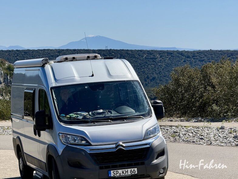 Ein silberner Citroën-Wohnwagen, geparkt auf einer Panoramastraße in der Ardeche, mit fernen Bergen und blauem Himmel im Hintergrund; grüne Sträucher und Bäume säumen die Landschaft. Unten rechts steht in weißer Schrift Hin-Fahren.