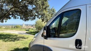 Ein weißes Wohnmobil ist auf einer Wiese in der Nähe eines Baumes geparkt und überblickt eine ländliche Landschaft mit einem Dorf, Feldern und entfernten Hügeln unter einem klaren blauen Himmel. In der Ecke steht der Schriftzug Hin-Fahren.