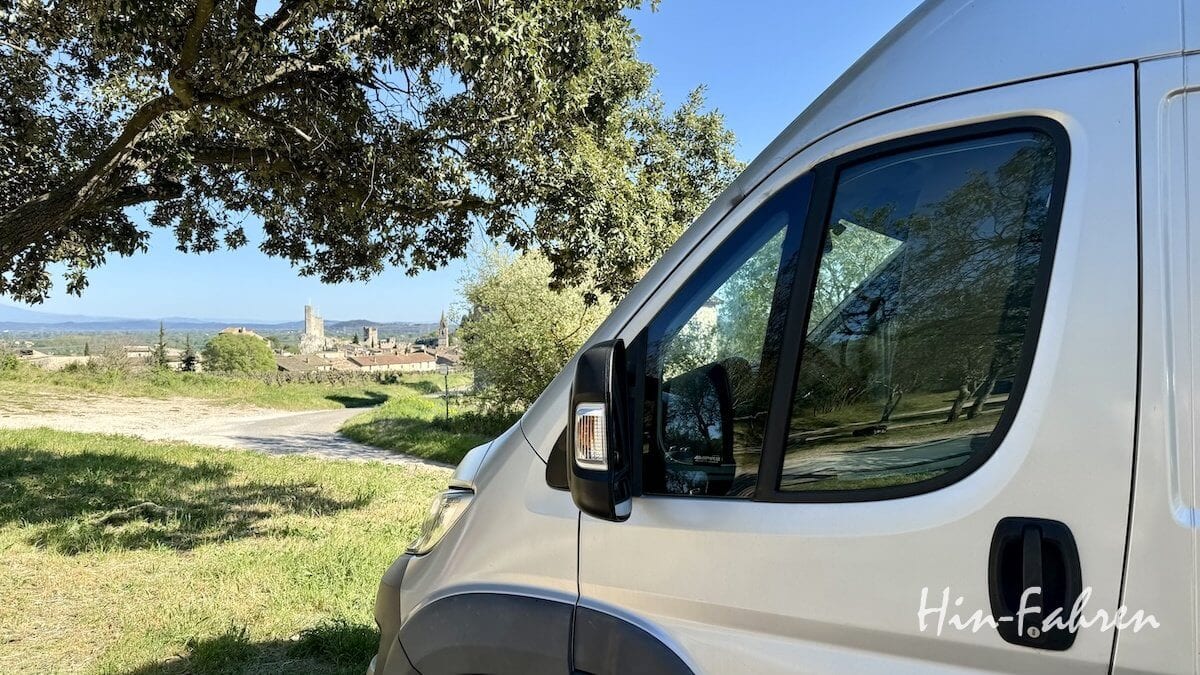 Ein weißes Wohnmobil ist auf einer Wiese in der Nähe eines Baumes geparkt und überblickt eine ländliche Landschaft mit einem Dorf, Feldern und entfernten Hügeln unter einem klaren blauen Himmel. In der Ecke steht der Schriftzug Hin-Fahren.