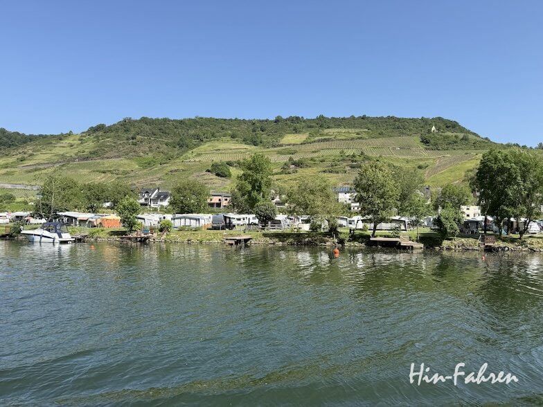 Ein ruhiger Fluss spiegelt Bäume, Häuser und Boote entlang seines Ufers in der Nähe des Campingplatzes Mosel wider, im Hintergrund sind grüne Hügel und Weinberge unter einem strahlend blauen Himmel zu sehen. In der rechten unteren Ecke steht in weißer Schrift „Hin-Fahren“.