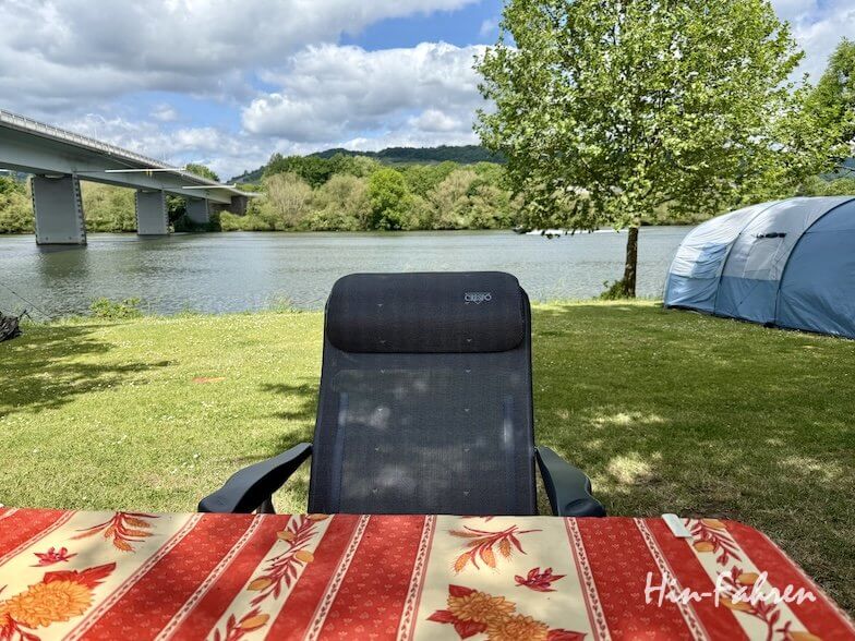 Auf dem Campingplatz Mosel steht ein Campingstuhl mit Blick auf den Fluss neben einem Tisch, der mit einer rot-gelb gemusterten Tischdecke gedeckt ist. Rechts steht ein blaues Zelt, links eine Brücke, im Hintergrund sind Bäume und Hügel unter einem teilweise bewölkten Himmel zu sehen.