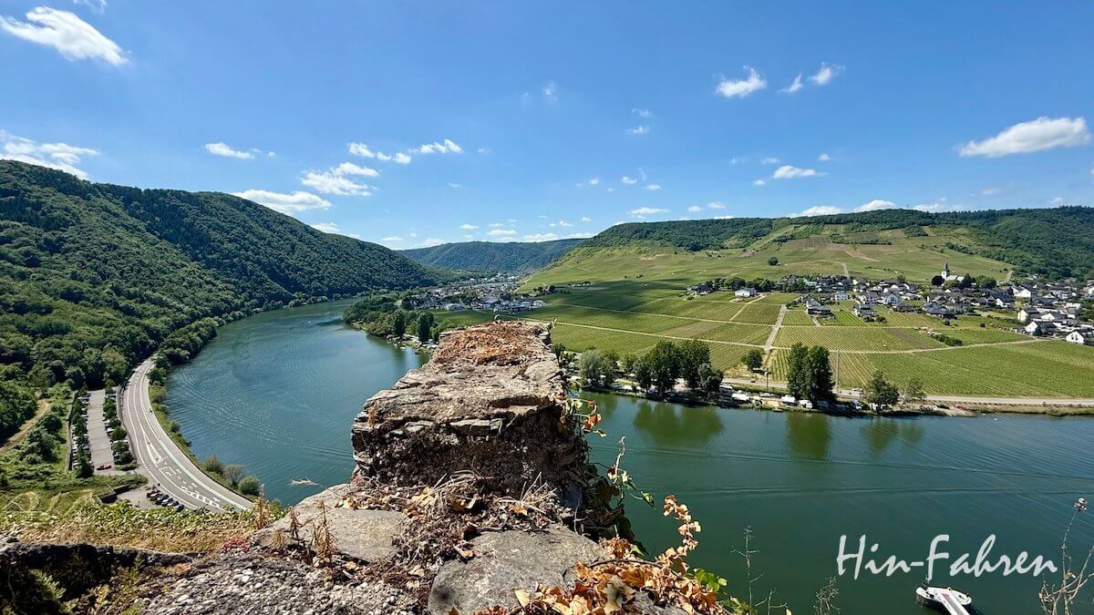 Blick von einem Felsvorsprung auf die sich schlängelnde Mosel, üppig grüne Hügel, Weinberge und ein kleines Dorf in der Nähe des Campingplatzes Mosel unter einem blauen Himmel mit vereinzelten Wolken. Unten rechts steht in Schreibschrift „Hin-Fahren“.