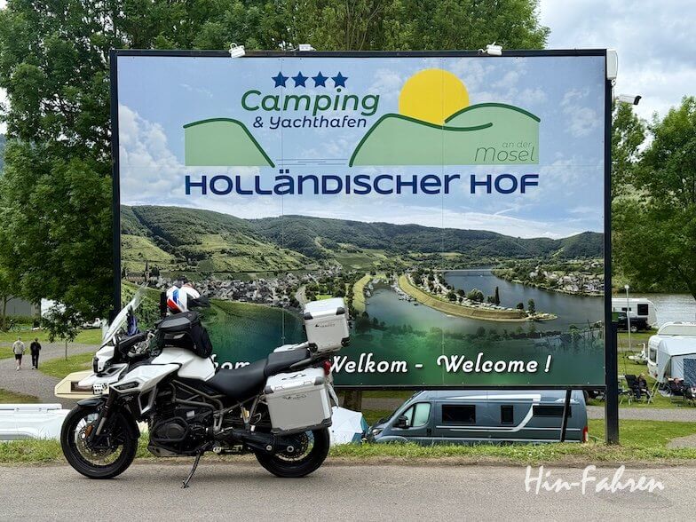 Vor einer großen Werbetafel des „Campingplatz Mosel“ mit der Aufschrift „Holländischer Hof“, auf der grüne Hügel, ein Fluss und die Sonne zu sehen sind, steht ein weißes Motorrad. Im Hintergrund sind Zelte und Bäume zu erkennen, die die malerische Lage des Campingplatzes unterstreichen.