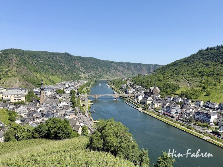 Ein Fluss schlängelt sich durch ein malerisches Tal, an dessen Ufern sich auf beiden Seiten der Campingplatz Mosel in der Nähe einer kleinen Stadt befindet. Eine Brücke überspannt den Fluss, und unter einem strahlend blauen Himmel erheben sich grüne Hügel. In der rechten unteren Ecke steht „Hin-Fahren“.