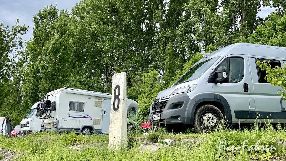 Titelbild Fun Facts Camping & Wohnmobil Zwei Wohnmobile und 1 Bulli stehen auf einer Wiese in der Nähe von Markierung 8, im Hintergrund sind hohe grüne Bäume zu sehen. Unter dem bewölkten Himmel sind eine kleine Blume und ein Fahrrad zu erkennen, das an einem der Wohnmobile befestigt ist – Wissenswertes: Camping in der Natur hebt die Stimmung und fördert die Kreativität!