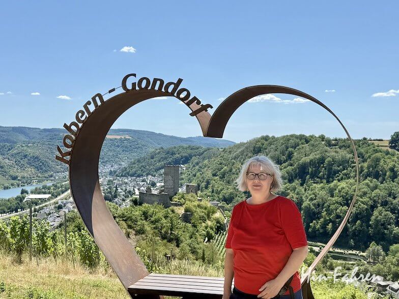Eine Frau in einem roten Hemd steht vor einem herzförmigen Metallrahmen mit der Aufschrift „Kobern-Gondorf“, im Hintergrund bietet sich unter strahlend blauem Himmel ein malerischer Ausblick auf Hügel, eine Burg und einen Fluss.