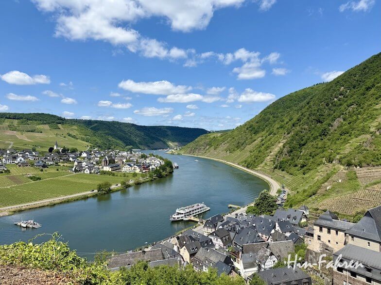 Ein malerischer Fluss schlängelt sich durch grüne Hügel und ein Dorf mit Häusern mit Schieferdächern in der Nähe des Campingplatzes Mosel. Ein weißes Boot gleitet auf dem Fluss dahin, während Weinberge die Hänge unter einem strahlend blauen Himmel mit vereinzelten Wolken bedecken.