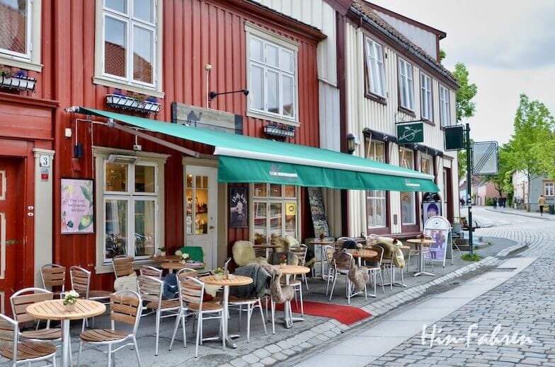 Ein Straßencafé mit Holztischen und -stühlen liegt an einer Kopfsteinpflasterstraße neben roten und beigen Holzgebäuden – ideal für alle, die Trondheim mit dem Wohnmobil erkunden. Eine grüne Markise spendet Schatten; üppig belaubte Bäume tragen zur friedlichen Atmosphäre bei.