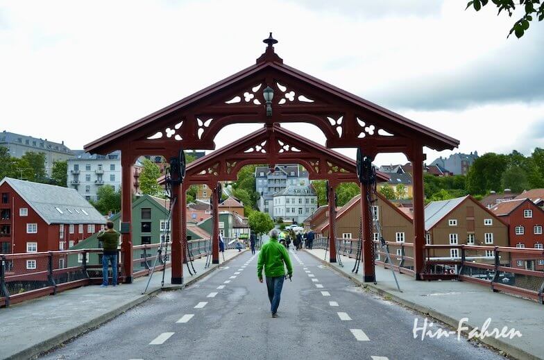 die alte Brücke Gamle Bybro in Trondheim: Eine Person spaziert mit dem Wohnmobil auf einer breiten Holzbrücke mit einem dekorativen roten Bogen in Trondheim, umgeben von bunten Häusern, üppigem Grün und bewölktem Himmel.