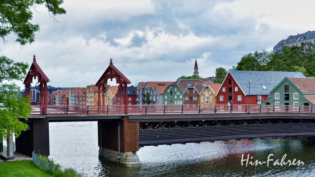 Blick auf die Alte Brücke und die bunten Holzhäuser der Altstadt von Trondheim. Eine historische Holzbrücke mit roten Bögen überspannt einen Fluss in Trondheim, wo unter einem bewölkten Himmel bunte Häuser das gegenüberliegende Ufer säumen.