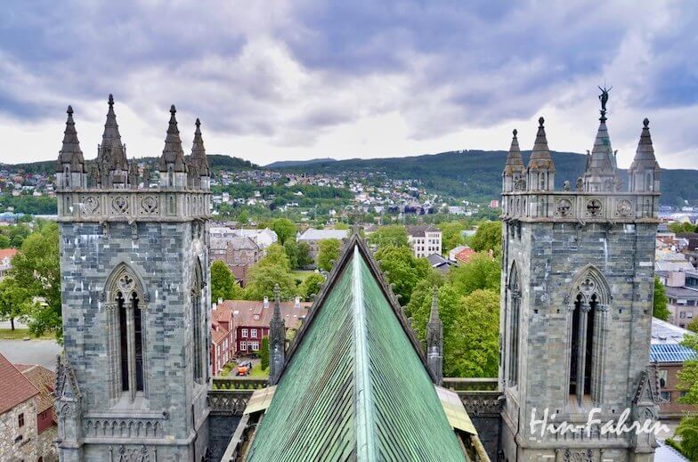 Luftaufnahme einer Kathedrale im gotischen Stil mit zwei Türmen in Trondheim, ideal für Erkundungstouren mit dem Wohnmobil, umgeben von grünen Bäumen, Gebäuden, Hügeln und einem bewölkten Himmel im Hintergrund.